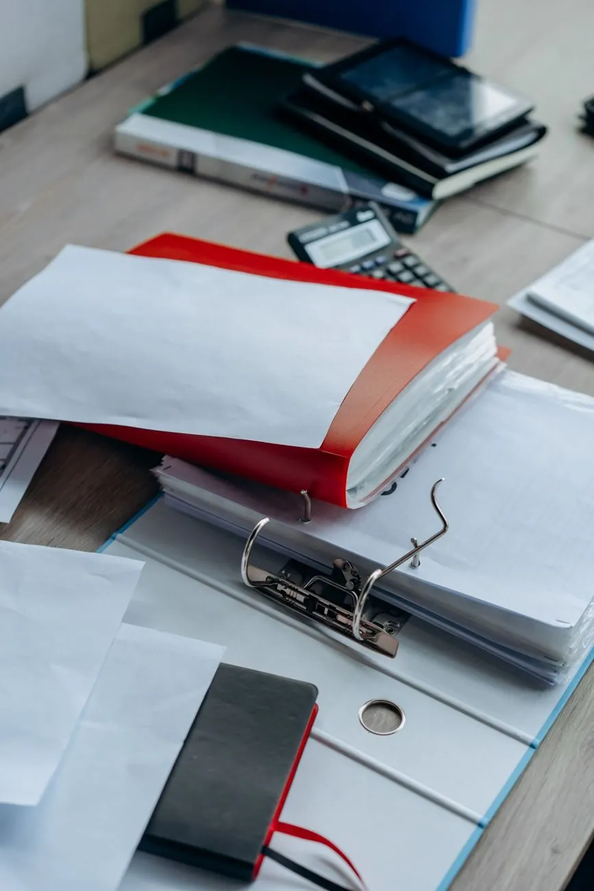 An open ring binder of papers with a red folder and a calculator on an office desk