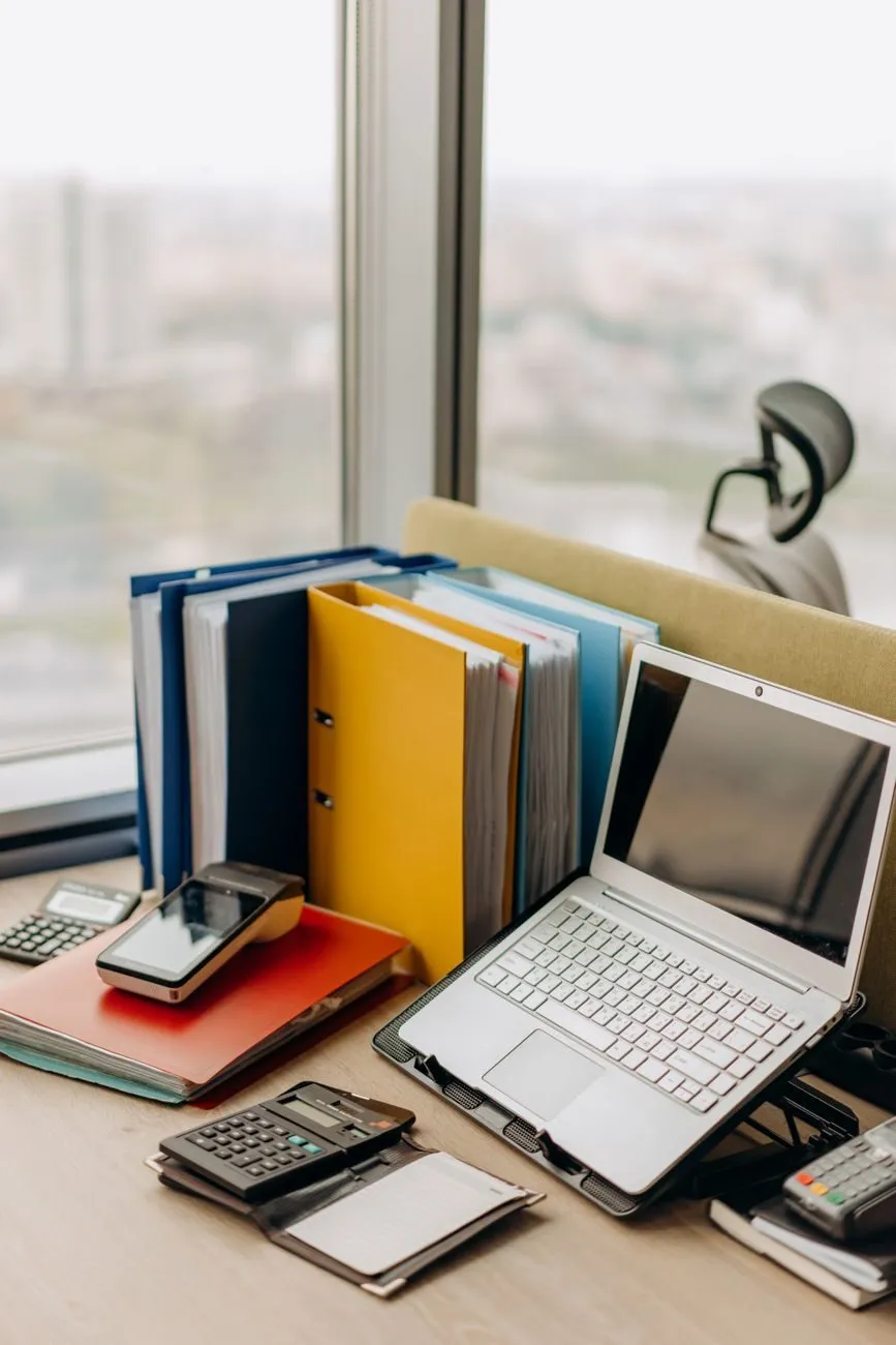 A desk by the window in the office, with binders, a laptop and calculators