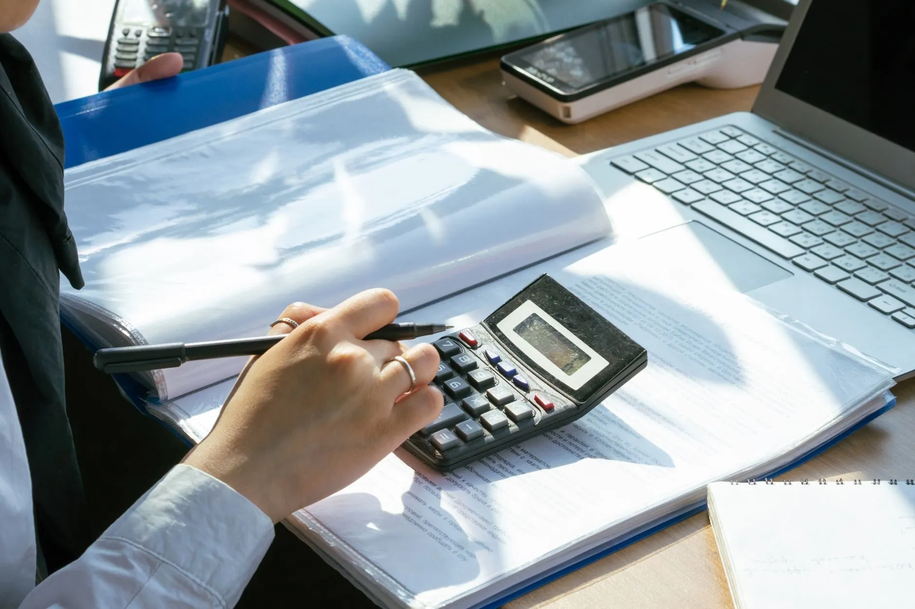 A hand working a calculator over a blue folder of invoices beside a laptop